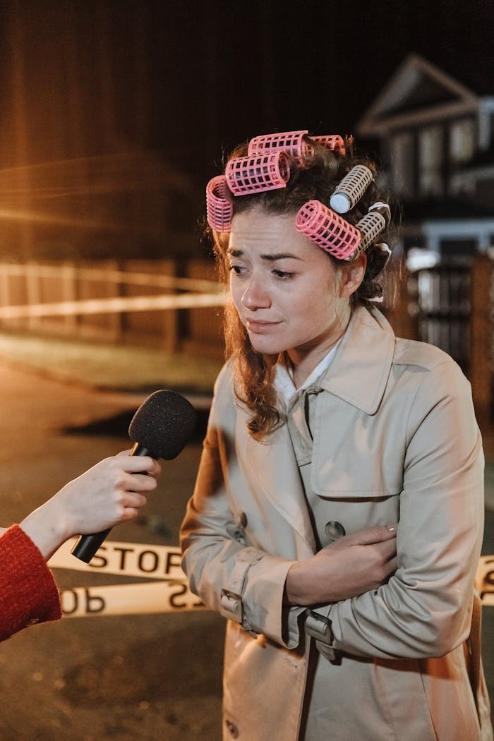 Woman with hair curlers and worried look being interviewed at a crime scene outdoors at night.