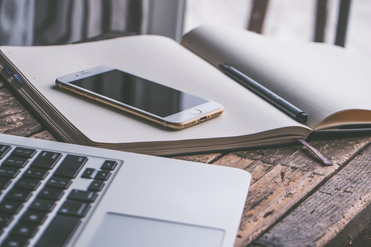 Open notebook with a smartphone and laptop on a wooden desk, creating a contemporary workspace vibe.